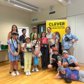 A group of children and adults posing together in a room with a wooden floor. Some children are holding bags and wearing hats. Behind them is a yellow sign with text, and there are boxes on a table to the left. Everyone is smiling.