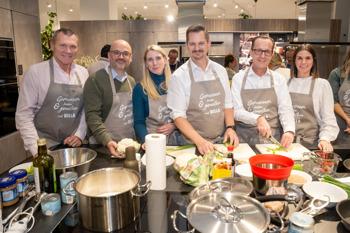 A group of six people wearing aprons stand around a kitchen counter preparing a meal. They are surrounded by cooking ingredients and tools, including pots, pans, and vegetables. The atmosphere is cheerful and cooperative.