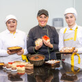 Three bakers in uniforms pose with a variety of baked goods, including cakes and pastries, in a kitchen setting.