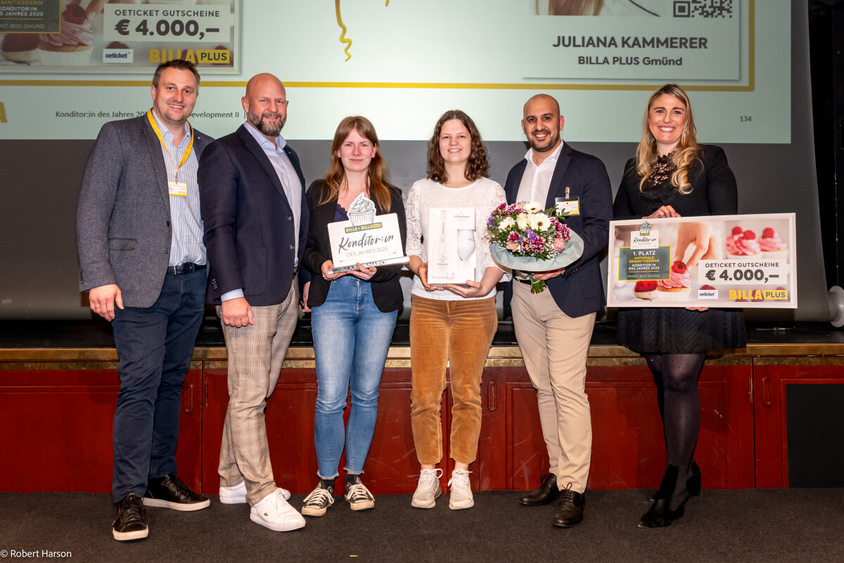 A group of six people stands on a stage, smiling. Three hold awards, including a framed certificate and a bouquet of flowers. In the background, a large presentation screen displays "Juliana Kammerer" and "Billa Plus" along with a gift certificate.