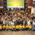 A large group of people gathered in a conference hall, some wearing yellow shirts, posing for a group photo in front of a screen displaying "nextbilgeneration Event 2024."