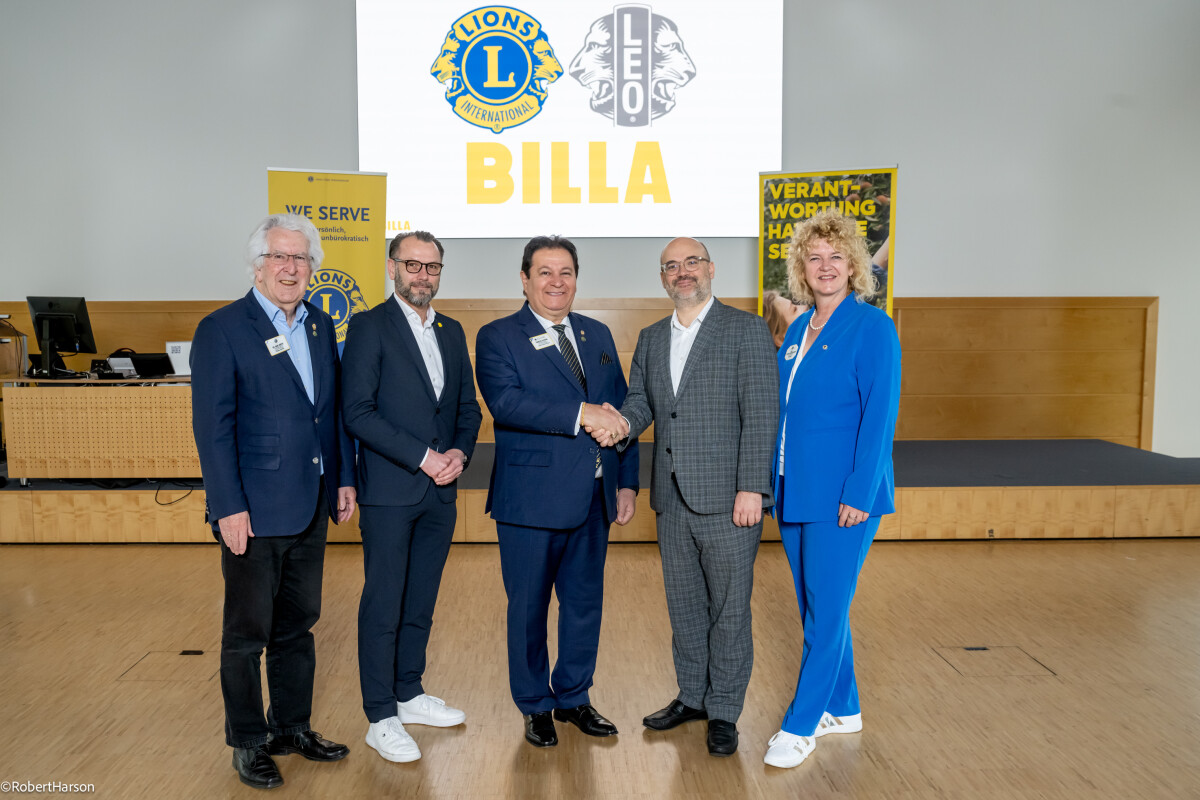 Five people in formal attire stand in a row, smiling, and shaking hands in a venue. A large screen behind them displays logos of Lions International, Leo, and Billa.