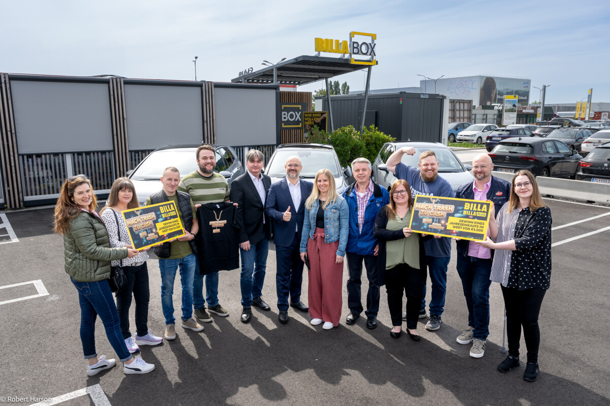 A group of people standing in a parking lot in front of a Billa Box, holding large yellow signs. They are smiling and posing for the camera. Some are giving thumbs up. The background features parked cars and the Billa branding.
