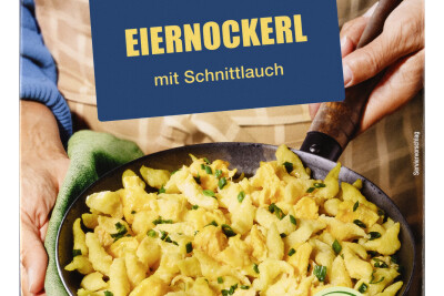 A person holding a bowl of Eiernockerl with chives. Packaging reads "Omis Schmankerl von Billa," highlighting the dish as vegetarian and protein-rich, with a quick 3.5-minute preparation time.