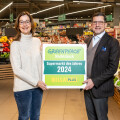 Two people standing in a supermarket holding a sign that reads, "Greenpeace Marktcheck Supermarkt des Jahres 2024 BILLA PLUS." They are smiling, surrounded by fruits and vegetables.