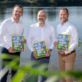 Three men in white shirts, standing by a lake, each holding a book titled "Wasser-Welt." They are smiling and surrounded by greenery, with the water and sky in the background.