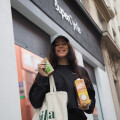 A woman in a black sweatshirt and a gray cap stands outside a shop, holding a canvas tote bag and two products: a green packaged item and a carton of billa bio oat milk. She is smiling and the shop has a modern facade.