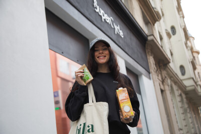 A woman in a black sweatshirt and a gray cap stands outside a shop, holding a canvas tote bag and two products: a green packaged item and a carton of billa bio oat milk. She is smiling and the shop has a modern facade.