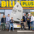 A group of people standing in front of a Billa Plus store, posing around a small sailboat with the number 1031. Some are casually dressed and two are holding award plaques. A poster with food images is on the right.