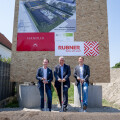 Three men in suits stand holding shovels at a groundbreaking ceremony. Behind them is a large sign with building plans and company logos, including Rubner and Handler. There is a pile of dirt in the foreground and construction equipment in the background.