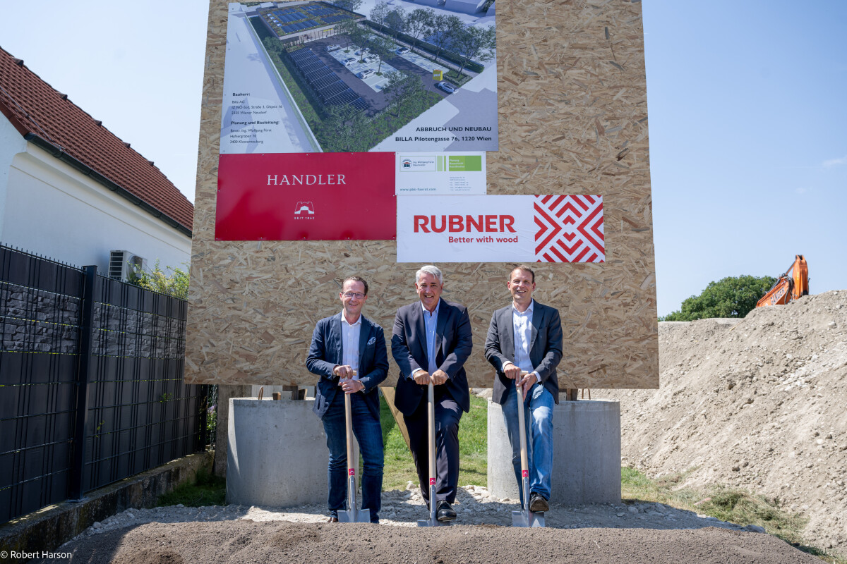 Three men in suits stand holding shovels at a groundbreaking ceremony. Behind them is a large sign with building plans and company logos, including Rubner and Handler. There is a pile of dirt in the foreground and construction equipment in the background.