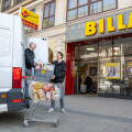 Two men unloading a delivery van and placing groceries into a shopping cart in front of a Billa supermarket. One man is handing over a box, and the other is standing with the cart, surrounded by a city street.