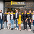 A group of nine people, wearing diverse styles of business and casual attire, stand smiling in front of a store with a "BILLA TO GO" sign above the entrance. They are positioned in a line, showcasing a friendly and professional atmosphere.