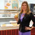 A woman in a black blazer and purple dress holding a sandwich stands in front of a counter displaying various baked goods. Signs advertise a product for 2.99 with labels indicating it is vegetarian and organic.