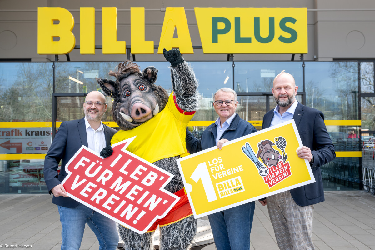 Three men stand outside a BILLA PLUS store with a boar mascot in a yellow and red outfit. The mascot holds a sign saying "Leb' für mein' Verein!" while one man holds another sign promoting "Los für Vereine" with sports icons. They are all smiling.