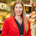 A woman with long blonde hair, wearing a red blazer and black top, is smiling and standing indoors. She is in front of shelves displaying various products, with bright, colorful signage in the background.