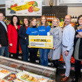 A group of people standing in a grocery store, holding a large check marked €27,905. Some people are wearing matching blue shirts with logos. The background includes various fruits, vegetables, and grocery items.