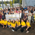 A large group of people gathered outdoors, posing for a photo. Several individuals in the front row are wearing bright yellow shirts. They are holding a colorful welcome sign with various handwritten messages. The setting is a wooden deck with greenery and wooden structures in the background. Everyone appears happy and engaged.