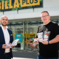 Two men standing outside a Billa Plus store, both holding packaged products and smiling. The man on the left is in a suit, and the man on the right is in a black T-shirt. The store entrance and signage are visible in the background.
