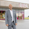 A man in a gray suit stands smiling in front of a BIPA store, with a modern exterior and glass doors. The background shows a parking area and a green field.