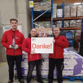 Three people wearing red jackets are standing in a warehouse. Two are holding decorative bottles, and the person in the middle is holding a sign that says "Danke!" They're standing in front of stacked goods on pallets.