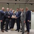 A group of eight people dressed in business attire stands in a grocery store aisle. They are holding a framed certificate or award together and smiling at the camera. Shelves stocked with various grocery items are visible in the background.