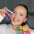 A person smiling and holding several colorful, cube-shaped lip balms in one hand, and two lip gloss tubes in the other. They have long hair pulled back and are wearing a light top. The background shows a bed and a light-colored wall.