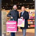 Two men standing in the entrance of a store, smiling and holding branded bags. One holds a large pink "Foodora" delivery bag, and the other holds a smaller shopping bag with German text. The store interior is visible in the background.
