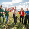 Five people standing in a green field on a sunny day, holding and examining plants. A sign with "ja! Natürlich" is in the middle. They appear relaxed and cheerful, dressed in casual clothing.