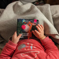 A child in a pink outfit holds a colorful book featuring a cartoon reindeer with a red nose. The book has the word "BIPA" on it, and the child is lying on a gray blanket.