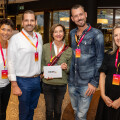 Five people smiling and posing together indoors, wearing name badges. The person in the center holds a REWE+ envelope. The group stands on a speckled floor with a modern, well-lit interior in the background.