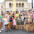 A group of people, including adults and children, smiling and making thumbs-up gestures in front of a historical building. They are gathered around a "Billa Corso Servicebike" and surrounded by brown paper bags.