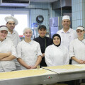 A group of seven people wearing chef uniforms stand smiling in a commercial kitchen. The kitchen features tiled walls and a large window. Dough and baking sheets are visible in the foreground.