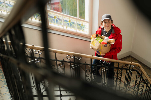 A person in a red jacket and cap stands on a staircase, holding a paper bag filled with groceries, including fresh produce. Natural light streams through a patterned window above.