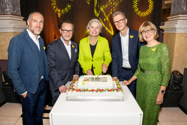 Five people stand around a decorated cake on a table, all wearing formal attire with yellow butterfly pins. The woman at the center, dressed in a lime green jacket, holds a knife to cut the cake as the others smile for the camera. The background is elegantly decorated with draped fabric and soft lighting.