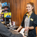A cashier wearing a yellow-collared uniform stands behind a counter, handing a receipt to a customer with long blonde hair. The background features shelves with products and a digital display.