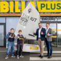 Two adults and two children smiling with thumbs up in front of a BILLA PLUS store. A sailboat with "TUA AUT 1031" on the sail is in the center.