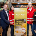 Four people stand indoors around a donation sign for a charity campaign. Two individuals wear red jackets, and two are in casual attire. The sign displays donation options of €5 and €10 to help families in need in Austria.