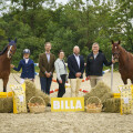 Five people standing in an outdoor arena with two horses. They are surrounded by hay bales and baskets of potatoes, with a "BILLA" sign in front. Trees are visible in the background.