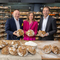 Three people smiling and holding loaves of bread in a bakery. They are standing in front of shelves filled with various breads, with more loaves and flour on the table in front of them.