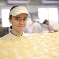 A person in a white uniform and cap is holding a tray filled with uncooked pasta pieces in a busy kitchen setting.