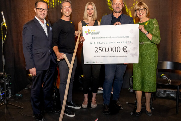 Five people standing on a stage holding a large check for 250,000 euros. One person is holding a large scythe. They are dressed in formal attire with a backdrop featuring bright light patterns.