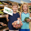 Two smiling children in a grocery store, holding produce wrapped in net bags. The boy holds a sign that says "Wunderlinge". They stand in front of a shelf filled with fruits and vegetables.