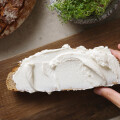 A hand holding a slice of bread generously spread with creamy white cheese, placed on a wooden board. Nearby are a loaf of rustic bread, a small bowl of salt, and a pot of fresh greenery.
