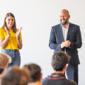 A woman in a yellow shirt claps while a man in a suit stands next to her, both facing an audience. The man is smiling, and people in the foreground are out of focus.