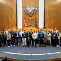 A group of people standing in a large, formal room with wooden paneling, a podium, and two flags, including the European Union flag. They are posing for a photo, wearing formal or business-casual attire.