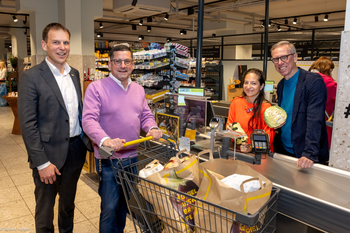 Four people are smiling near a supermarket checkout counter. One man is pushing a shopping cart filled with groceries in paper bags. A woman and another man are on the other side of the counter, holding a payment terminal and a cabbage. A fourth man in a suit stands nearby.