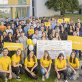 A group of people, including young adults and adults, gather outside smiling. Some are wearing yellow shirts. They're holding signs with text like "#nexbillageneration" and "Willkommenstage." Balloons and a large poster are present. Buildings are in the background.