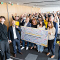 A large group of people posing for a selfie at an indoor event. Many are holding up a large, colorful sign with text and signatures. The room has wooden walls and some attendees are wearing yellow shirts. People are smiling and waving.