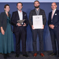 Four people standing in front of a blue backdrop. Two men in the center are holding an award and a certificate. They are flanked by a woman on the left and a man on the right, all dressed in formal attire.